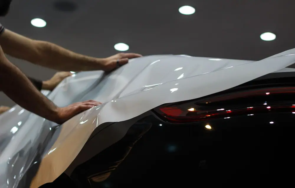 Hands laying a sheet of white film down over a curved body panel, working it into the shape
